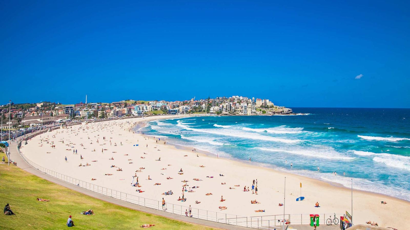 Phong cảnh toàn cảnh bãi biển Bondi nổi tiếng tại Sydney với bãi cát trắng mịn, làn nước biển xanh ngắt và các tòa nhà ven bờ dưới bầu trời trong xanh.